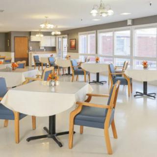 A bright and spacious dining area with neatly arranged tables and chairs, featuring large windows and overhead lighting.