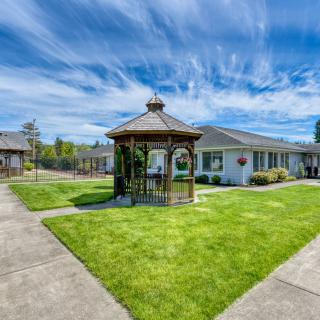 A picturesque gazebo stands in the center of a well-kept lawn, surrounded by pathways and residential buildings under a partly cloudy sky.