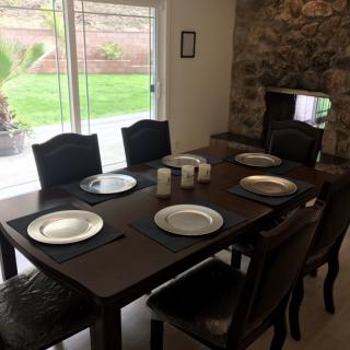 A dining room with a wooden table set for six, featuring plates and chairs, next to a large window and stone fireplace.
