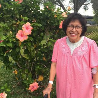 A woman in a pink dress smiles while standing next to a hibiscus bush in a garden.