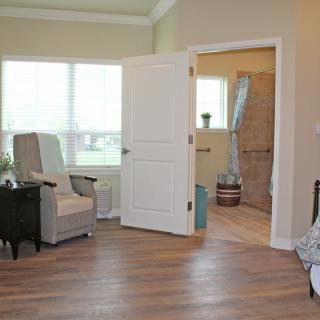 A cozy living room with a chair, open door leading to a bathroom, and natural light from a large window.