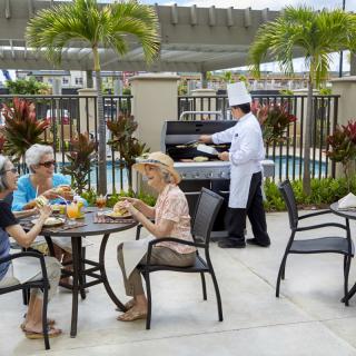 A group of people enjoying a meal outdoors while a chef cooks on a barbecue grill by the poolside.