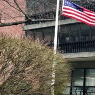 An American flag waves on a flagpole in front of a building, surrounded by trees.