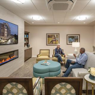 A cozy lounge area with two people having a conversation, featuring comfortable seating, a fireplace, and a wall-mounted television.