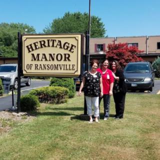 A group of people standing in front of the Heritage Manor of Ransomville sign.