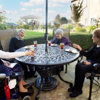 A group of elderly women enjoying drinks and conversation at an outdoor table in a garden setting.