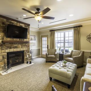 A cozy living room with comfortable seating and a stone fireplace, featuring a ceiling fan and large windows.