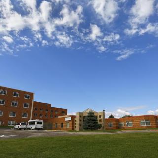 A view of a large building complex under a blue sky with scattered clouds, surrounded by grass and featuring a parked vehicle.