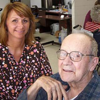 A nurse smiles while assisting an elderly man wearing glasses in a caring environment.