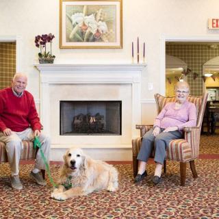 An elderly couple sitting by a fireplace with their dog in a comfortable living room.