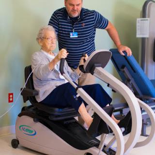 An elderly woman engages in a fitness session with the help of a personal trainer, using specialized exercise equipment for rehabilitation.