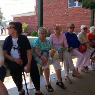 A group of elderly women sitting on a bench outdoors on a sunny day.