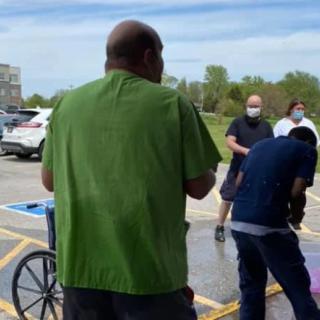 People gathered in a parking lot, some wearing masks, with a wheelchair visible.