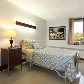 A clean and well-lit hospital room featuring a single bed with patterned bedding, a lamp on the nightstand, a framed artwork on the wall, and a large window providing natural light.