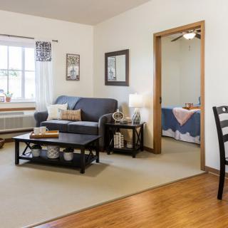 A cozy living room with soft lighting, featuring a sofa and coffee table, adjacent to a bedroom.