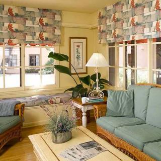 A cozy living room featuring a wicker sofa set with light green cushions, a wooden coffee table, large windows with patterned curtains, and decorative plants.