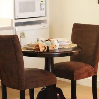 A small kitchen area with a dining table set for two, featuring brown upholstered chairs and a microwave in the background.
