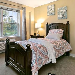 A cozy bedroom with a single bed, adorned with floral bedding and a soft throw, beside a window and a nightstand with a lamp.