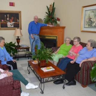 A group of seniors socializing in a comfortable living room setting with plants and artwork.