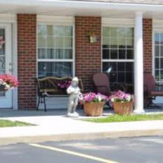A cozy patio area in front of a brick building with benches, chairs, flower pots, and a small statue.
