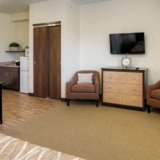 A spacious hotel room featuring a kitchenette, two armchairs, a dresser, and a wall-mounted TV.