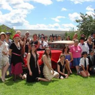 A group of people dressed in 1920s-themed costumes pose for a photo outdoors beside a classic car.