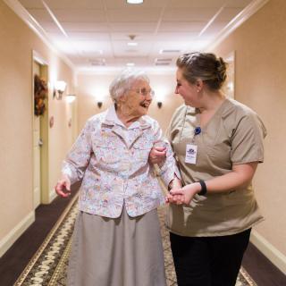 A caregiver assists an elderly woman as they walk together down a hallway.