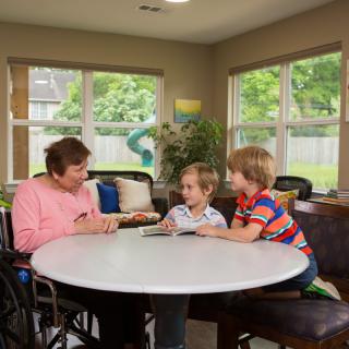 An elderly person and two children sitting at a table, engaging in a reading activity indoors.