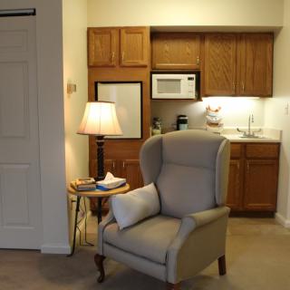 A cozy living area with a comfortable armchair and a lamp, adjacent to a small kitchenette.
