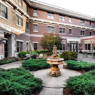 A charming courtyard featuring a central fountain surrounded by lush greenery and outdoor seating, set against a brick building backdrop.