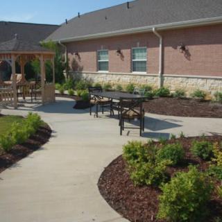 A landscaped outdoor area featuring a patio with seating, a pergola, and a walkway surrounded by shrubs and mulch in front of a building.