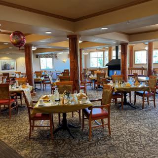 A cozy dining area with neatly arranged tables and chairs, featuring warm decor and ample natural light from large windows.