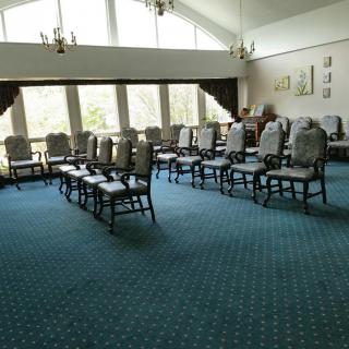 An empty room with rows of chairs arranged on a carpeted floor, featuring large windows and chandeliers.