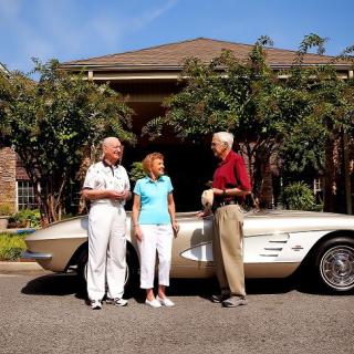 Three people standing beside a vintage car in front of a house.