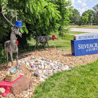 Metal deer sculptures wearing hats and ties are displayed near a sign for Silvercone at Lakewood amidst landscaped stones and greenery.