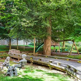 A tranquil playground surrounded by lush greenery, featuring a swing set and statues under the shade of large trees.