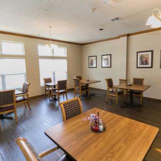 A cozy dining room with several tables and chairs, adorned with art on the walls and well-lit by large windows.