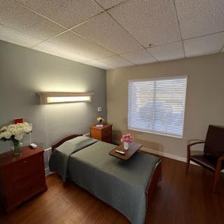 A cozy hospital room featuring a bed, bedside table, and a chair, with natural light coming from the window.