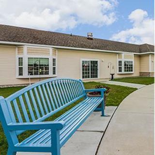 A blue bench sits by a pathway in front of a beige building under a partly cloudy sky.