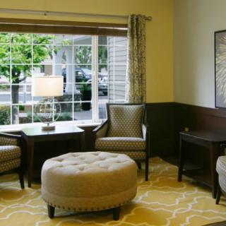 A cozy living room with upholstered chairs, a round ottoman, and a table lamp near a large window.