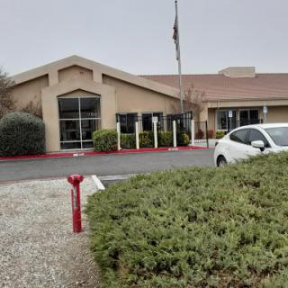 A view of a single-story building with a parking lot and greenery in the foreground, set against a cloudy sky.
