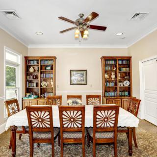 A well-furnished dining room with a large table surrounded by chairs, flanked by bookshelves and lit by a ceiling fan.
