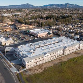 Aerial view of a suburban commercial building under construction, surrounded by a parking lot and residential neighborhood with mountains in the background.