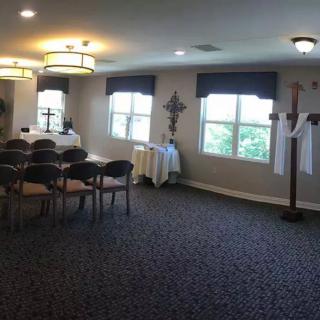 A small chapel room with chairs arranged in rows, featuring windows and a cross on the wall.