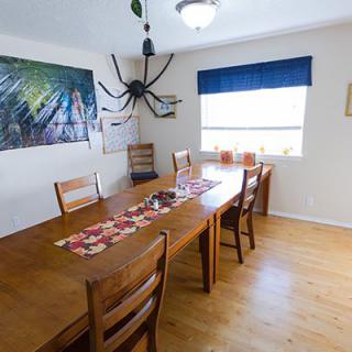 A dining room with a wooden table and chairs, featuring wall art and an oversized spider decoration hanging from the ceiling.