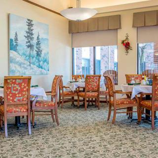 A cozy dining room with elegantly upholstered chairs and tables, featuring artwork and natural lighting from large windows.
