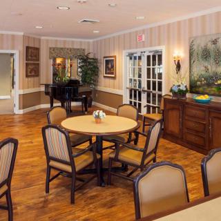 A cozy dining area with round tables and cushioned chairs on a wooden floor, featuring wall art and a piano in the background.