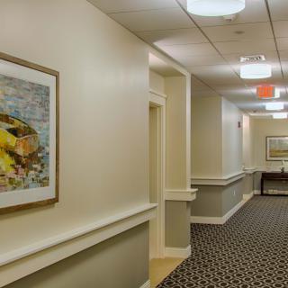 A well-lit hallway with framed artwork on the wall and patterned carpet flooring.