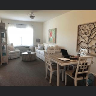 A cozy living room with a dining table and laptop setup, featuring neutral-toned furniture and natural lighting.