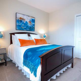 A neatly organized bedroom featuring a wooden bed with colorful pillows and a blue throw blanket, complemented by wall art and bedside lamps.